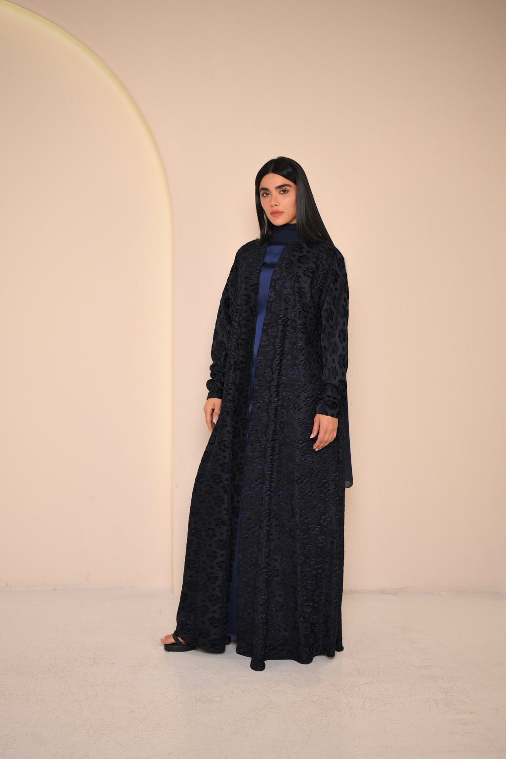Woman wearing a black abaya with a blue top against a beige background